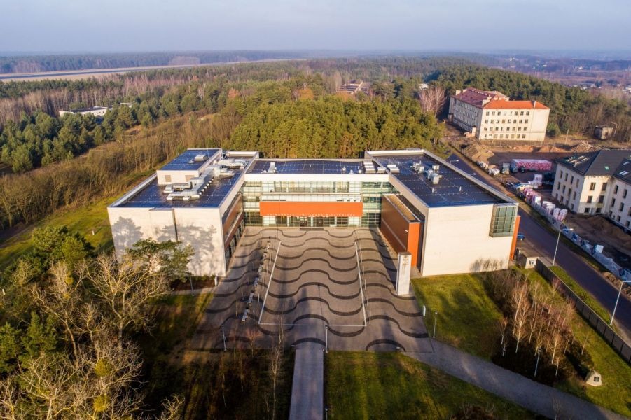 Widok z lotu ptaka na nowoczesny, duży budynek w kształcie litery U, otoczony lasem. Przed wejściem znajduje się plac z falistym wzorem nawierzchni, a wokół widać drzewa i kilka innych budynków.