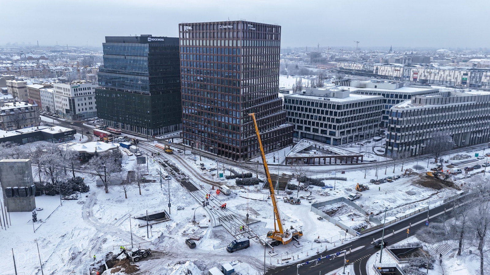 Widok z lotu ptaka na remonowane ulice. W centralnej części ujęcia widać dzwig i maszyny remontowe. W tle wysokie budynki i kamienice - panorama miasta. Wszystko w zimowej scenerii, przysypane śniegiem.