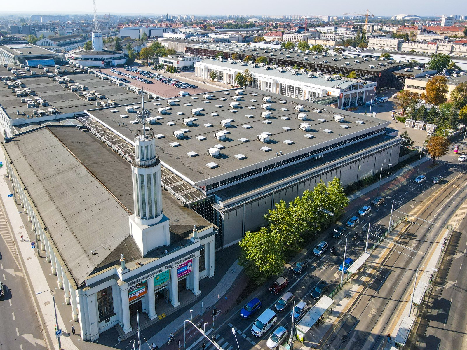 NetZero Energy i H2POLAND to wydarzenie, które skupia kluczowe tematy związane z przyszłością energetyki i neutralnością klimatyczną Widok z lotu ptaka na rozległy kompleks budynków, przypominający centrum wystawowe lub konferencyjne. Obiekty posiadają płaskie dachy z licznymi świetlikami i urządzeniami wentylacyjnymi. Jeden z budynków wyróżnia się klasyczną architekturą z kolumnami przy wejściu oraz wieżą ze spiczastą iglicą. W okolicy znajdują się parkingi, ulice z samochodami oraz inne budynki.