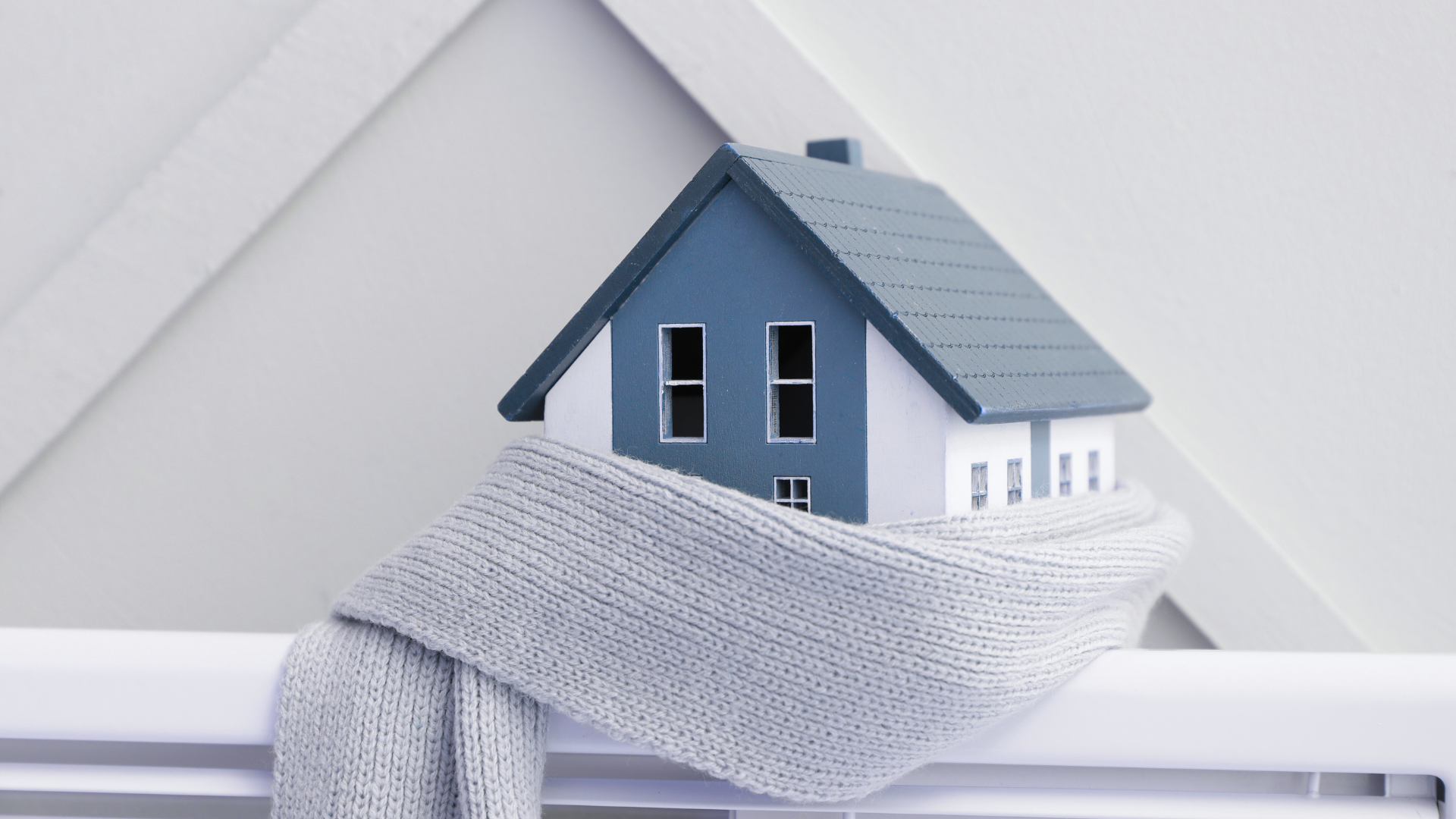 Zdjęcia w chłodnych kolorach szarym, niebieskim i białym przedatawia drewnany domek owinięty w wełniany szal umieszczony na grzejniku