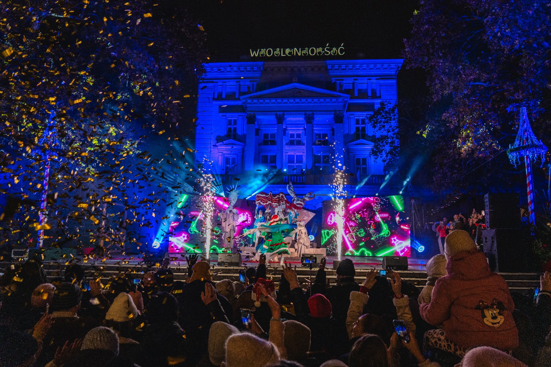 Zdjęcie przedstawia artysów na scenie i podświetlony budynek Arkadii.