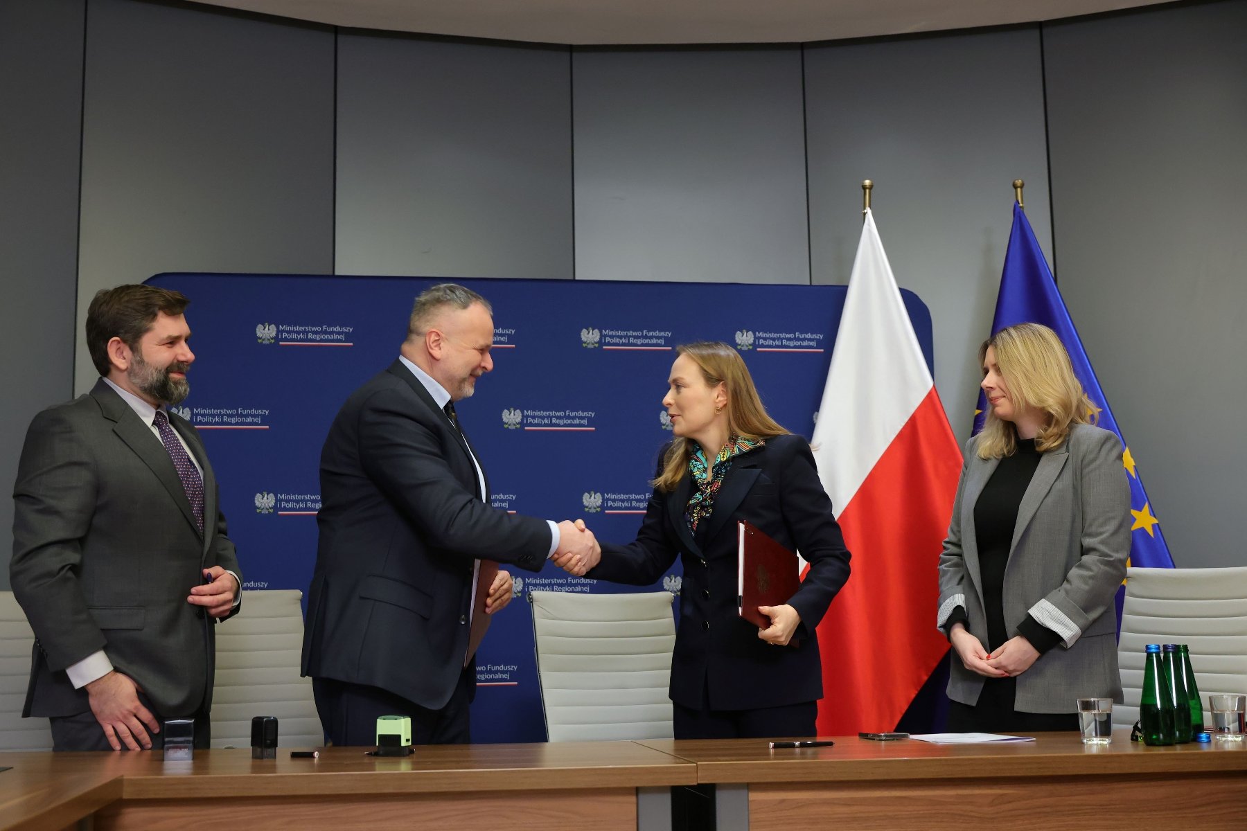 3 lutego w siedzibie Ministerstwa Funduszu i Polityki Regionalnej podpisano umowy o dofinansowanie projektów wybranych w konkursie na rozwój Społecznych Agencji Najmu (SAN)/ fot. ZKZL Zdjęcie przedstawia czwórkę ludzi - dwóch mężczyzn i dwie kobiety - stojących przy stole. Mężczyzna i kobieta znajdujący się pośrodku podają sobie ręce. W tle widać flagi Polski i Unii Europejskiej oraz ściankę z nazwą ministerstwa.