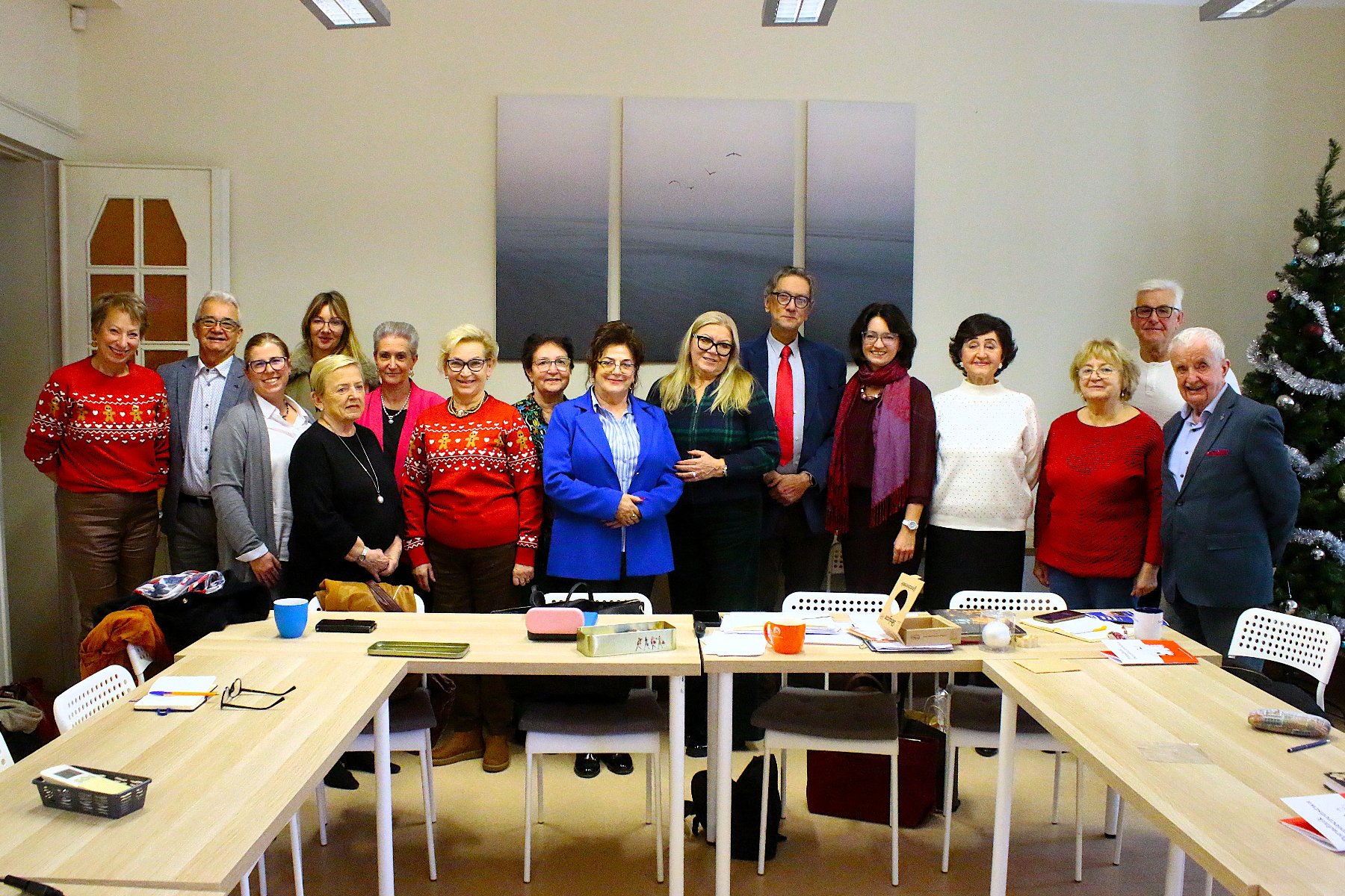 Zdjęcie przedstawia grupę seniorek i seniorów, którzy stoją przed stołami