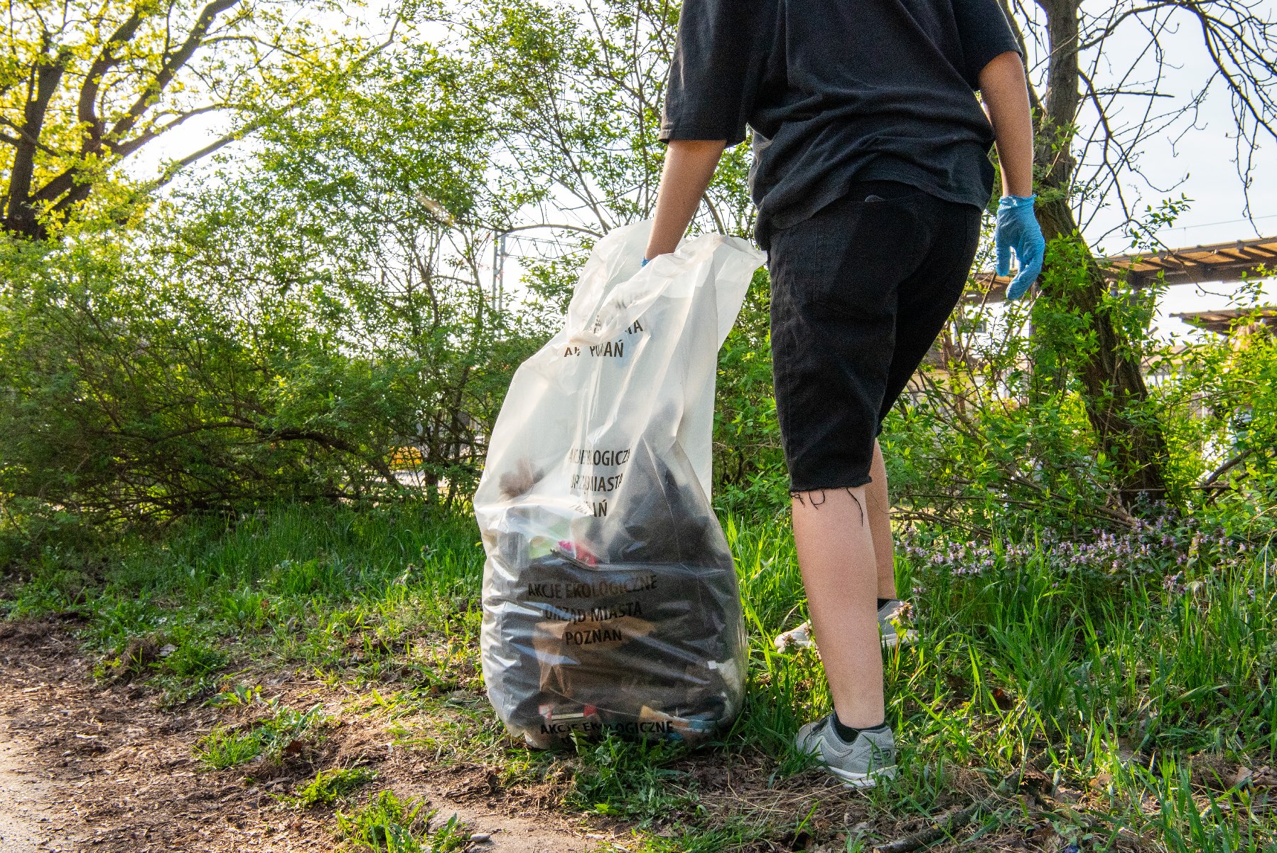 Podczas tegorocznych Wiosennych Porządków poznaniacy i poznanianki znaleźli wiele "skarbów". Łącznie zebrano 39 ton odpadów Zdjęcie przedstawia nogi osoby sprzatającej, która w ręce trzyma worek ze śmieciami. Obok widać zieleń, drzewa i krzewy.
