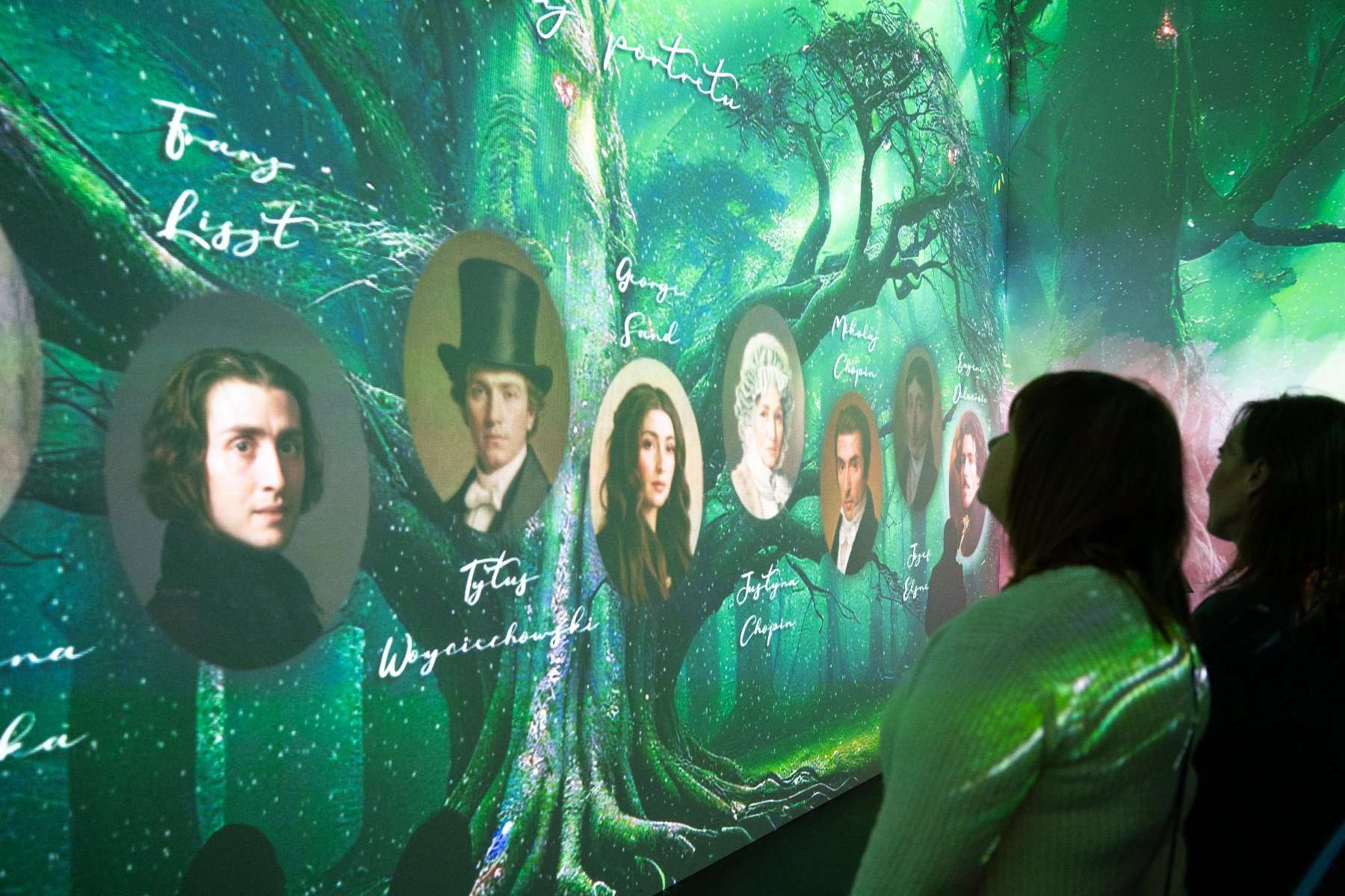 Zdjęcie przedstawia obraz wyświetlany na scianie, przedstawiajacy pięciolinie z nutami, oraz ludzi, którzy go oglądają i robią zdjęcia telefonami.