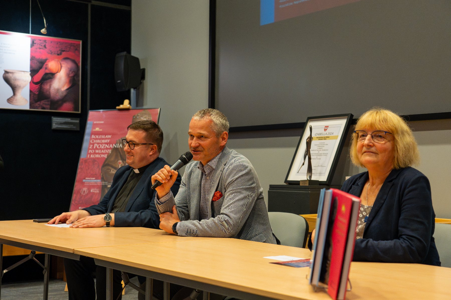 Zdjęcie przedstawia plakat z nazwą wystawy i wizerunkiem Chrobrego stojący przed salą, w której odbywa się konferencja prasowa.