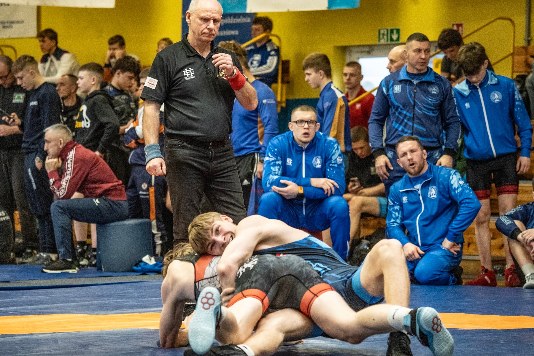 Międzynarodowy Turniej Zapaśniczy w Poznaniu, fot. Maciej Kula - 2 zawodnicy leżą na macie, w tle widać sędziego, przyglądających się trenerów oraz innych widzów na trybunach