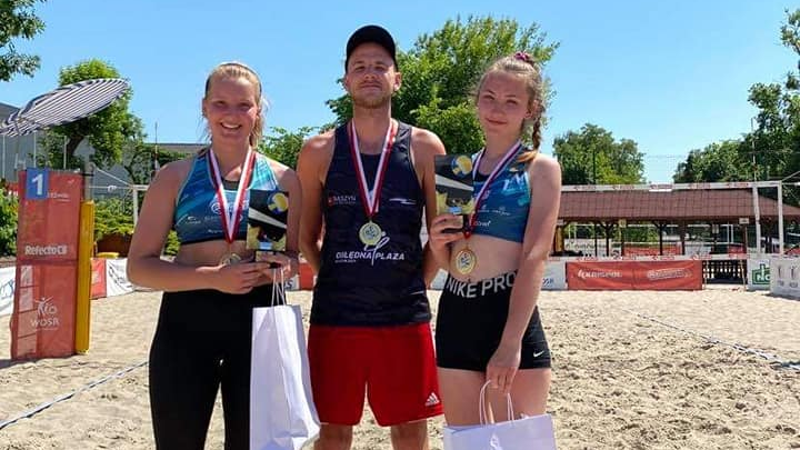 dwie zawodniczki oraz zawodnik stojący pośrodku na tle boiska do siatkówki plażowej - grafika artykułu