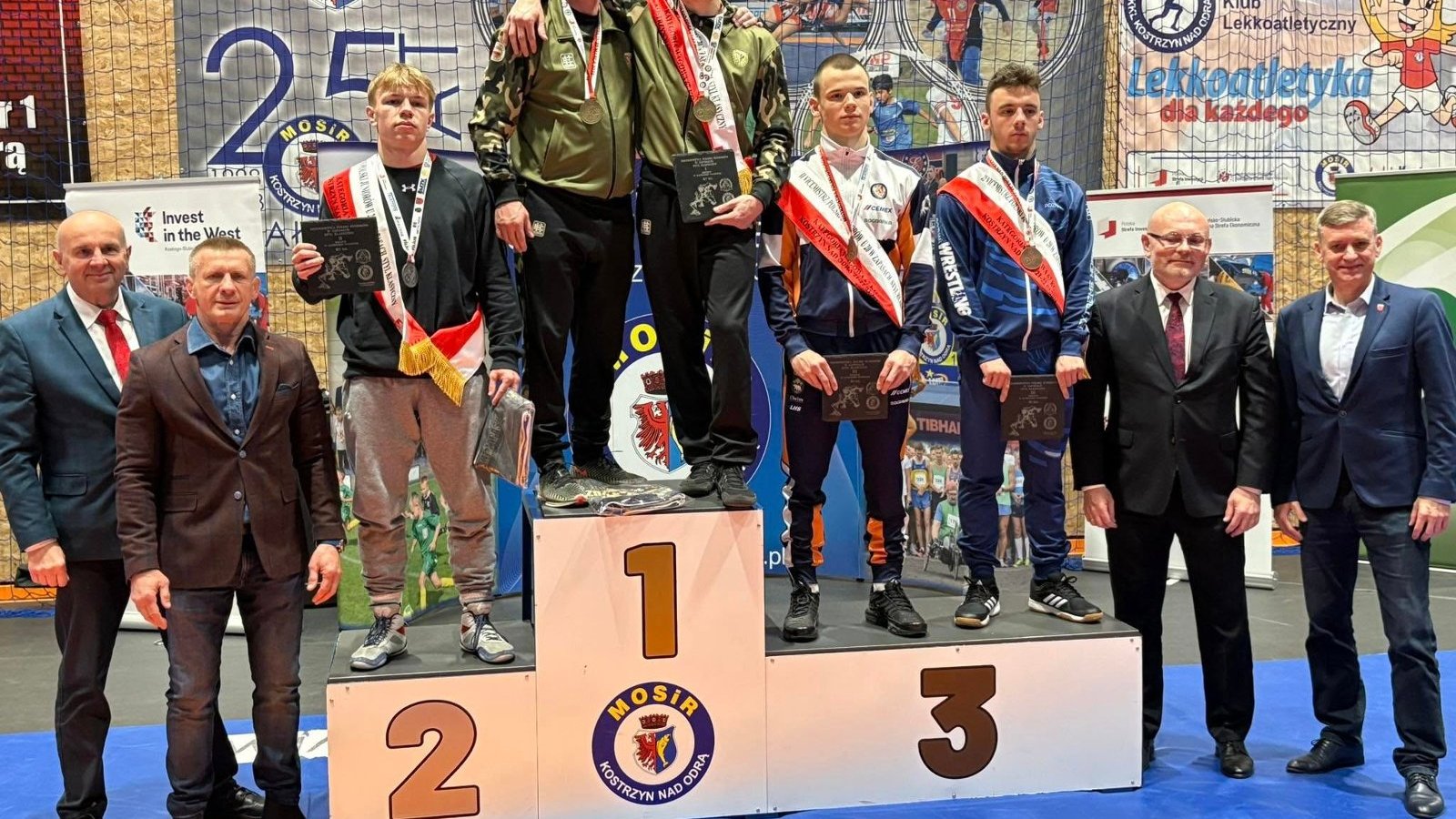 Zawodnik Sobieskiego Poznań na podium MP juniorów w zapasach, fot. Anna Zysk - na najwyższym stopniu podium stoi mężczyzna ze swoim trenerem w bluzie w kolorze moro. Na drugim stopniu podium stoi jeden zawodnik, a na trzecim dwóch zawodników. Wszyscy mają medale i statuetki. Obok podium stoi 4 oficjeli