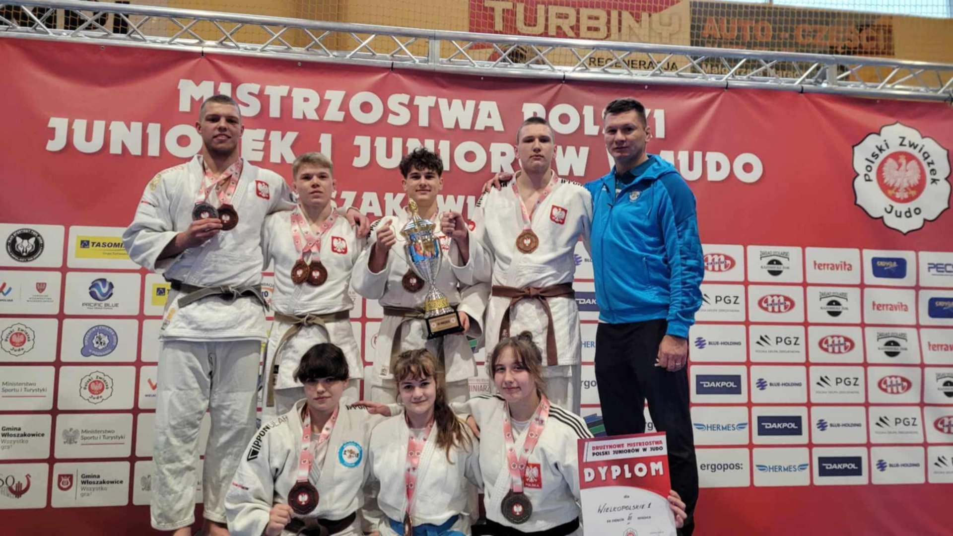 Zawodnicy Akademii Judo na MP juniorów w judo we Włoszakowicach, fot. Akademia Judo - na podium siedmioro zawodników z medalami i pcuahrami za 3. miejsce w drużynie razem ze swoim trenerem. Wszyscy zawodnicy stoją w kimono, a trener w niebiesko-czarnym dresie