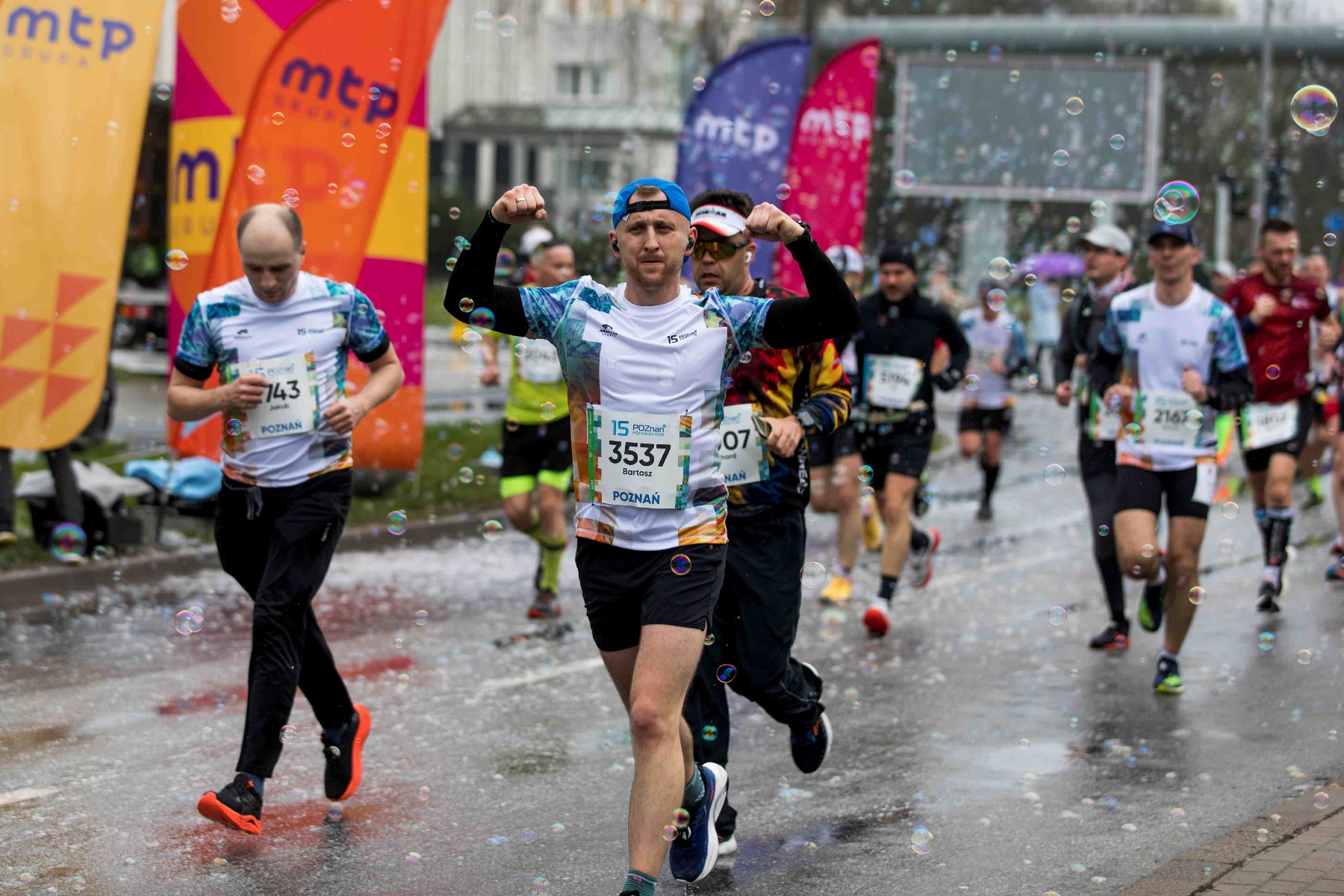 Poznań Półmaraton, fot. Agnieszka Weber-Ciereszko Poznań Półmaraton, fot. Agnieszka Weber-Ciereszko - biegacz w trakcie biegu podnosi ręce w górę, a w tle widać innych biegaczy, którzy biegną wśród baniek mydlanych.