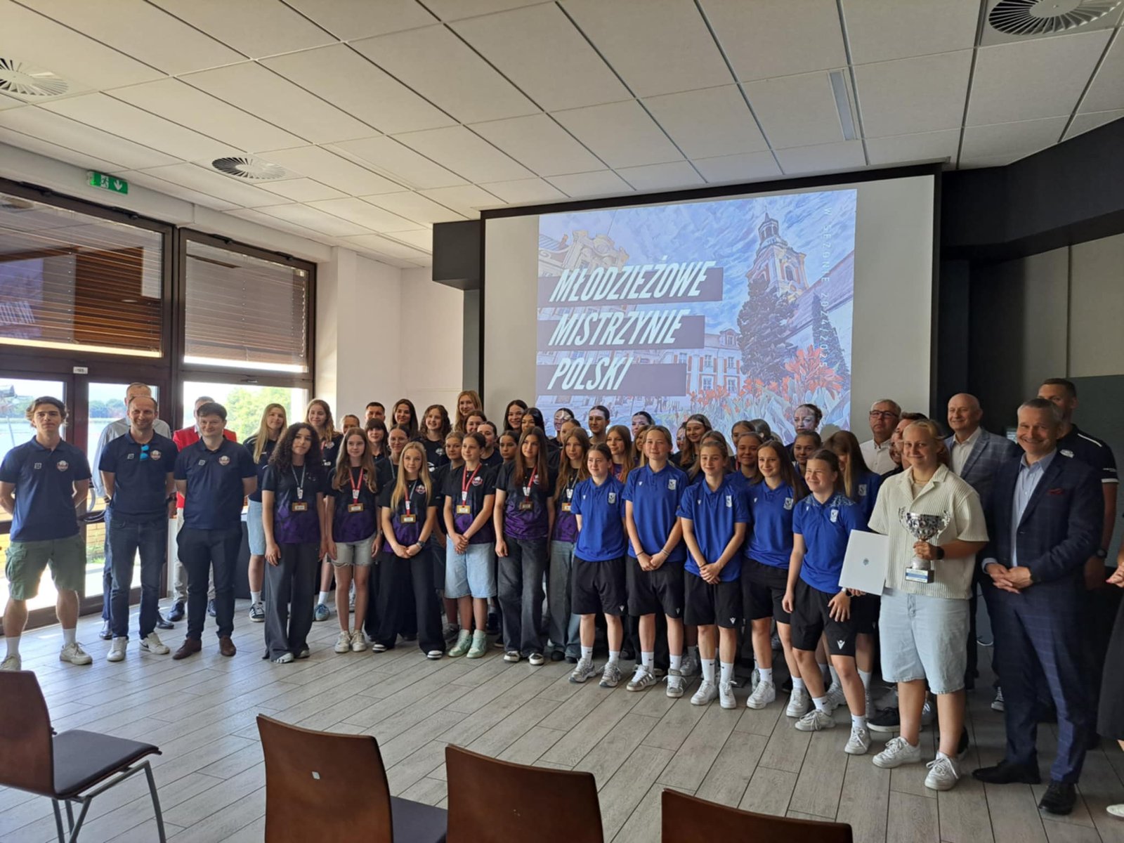 Spotkanie z Młodzieżowymi Mistrzami Polski Spotkanie z Młodzieżowymi Mistrzami Polski - kilkadziesiąt w sportowych strojach klubowych stoi na tle prezenacji z medalami, dyplomami i pucharami