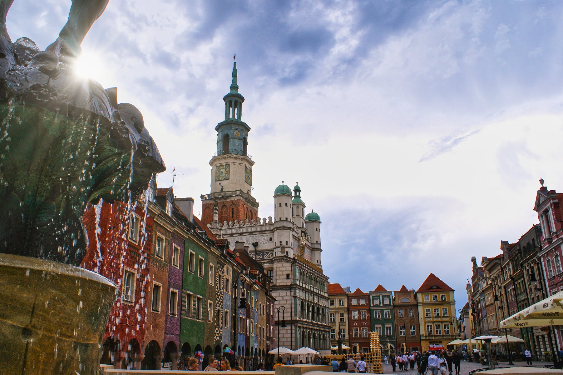 Stary Rynek w Poznaniu z żabiej perspektywy. W narożniku widać fontannę, woda pryska w promieniach słonecznych.
