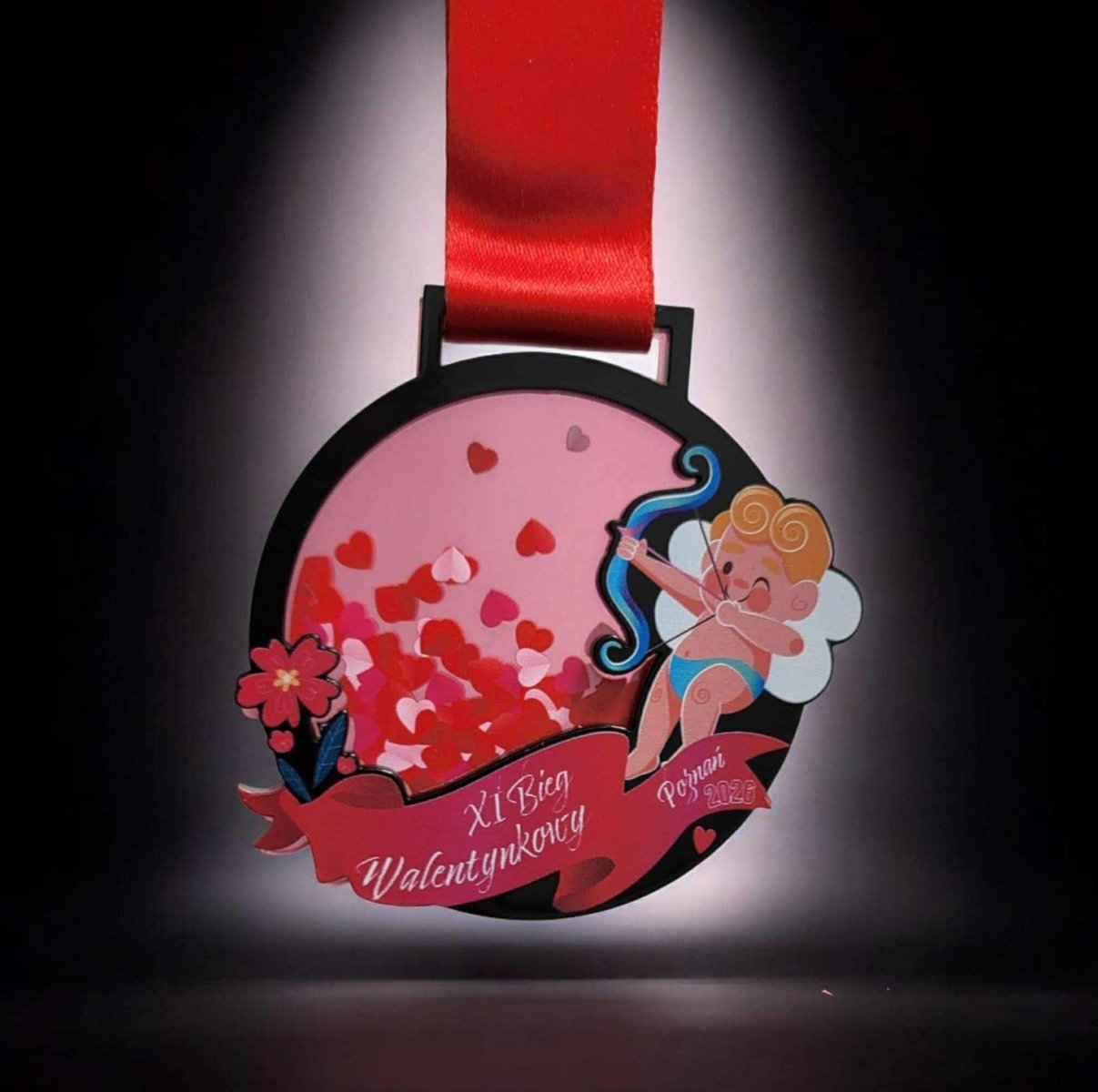 Wizualizacja medalu XI Biegu Walentynkowego - na ciemnym tle jest medal z wizerunkiem amorka, a także małymi czerwonymi sercami na różowym tle