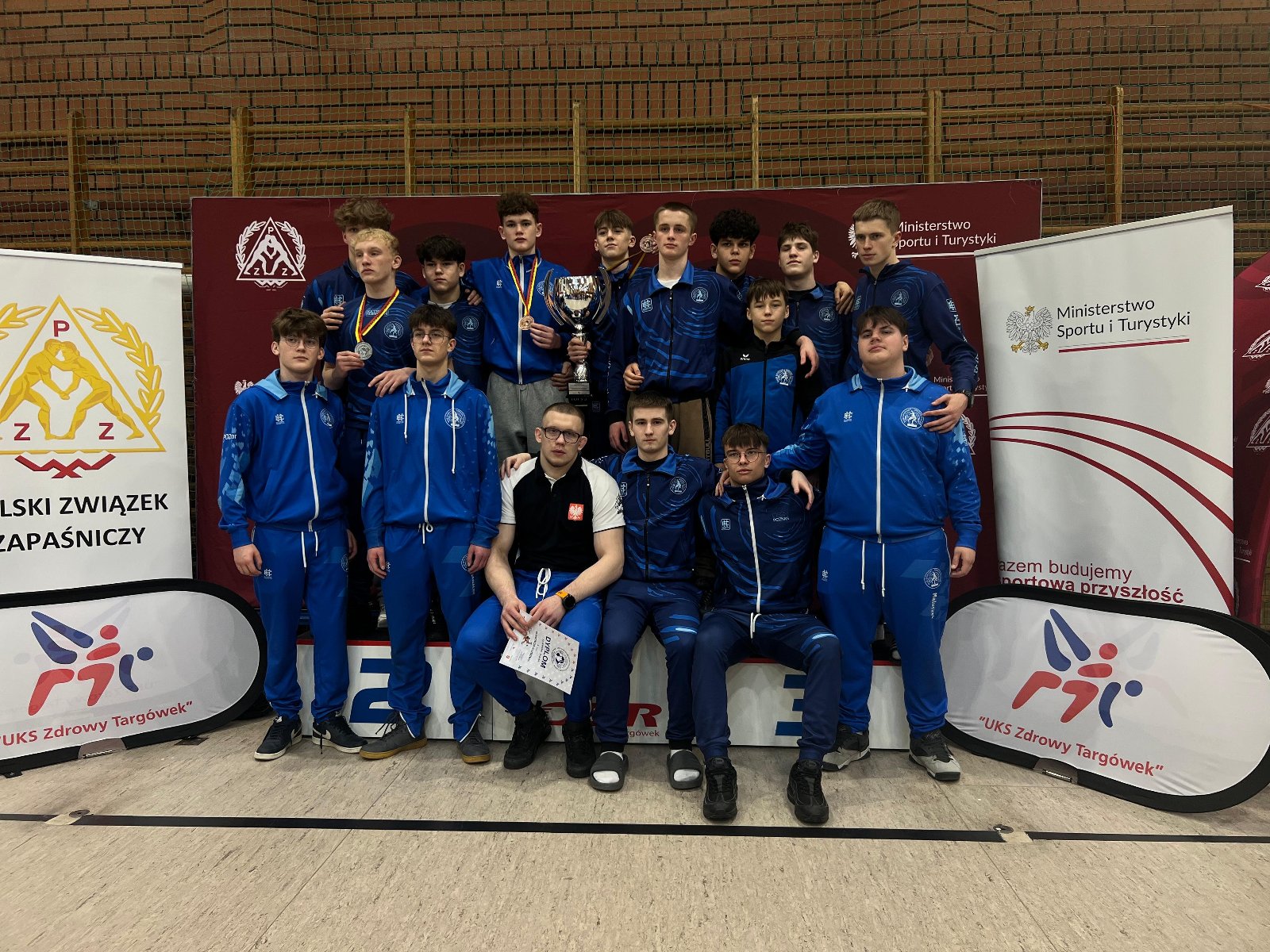 Zapaśnicy Sobieskiego na Pucharze Polski Kadetów w Warszawie, fot. Sobieski Poznań - na podium stoi kilkunastu zawodników w niebieskich dresach klubowych. Jeden z nich trzyma puchar, inny dyplom, a trzej mają medale na szyi