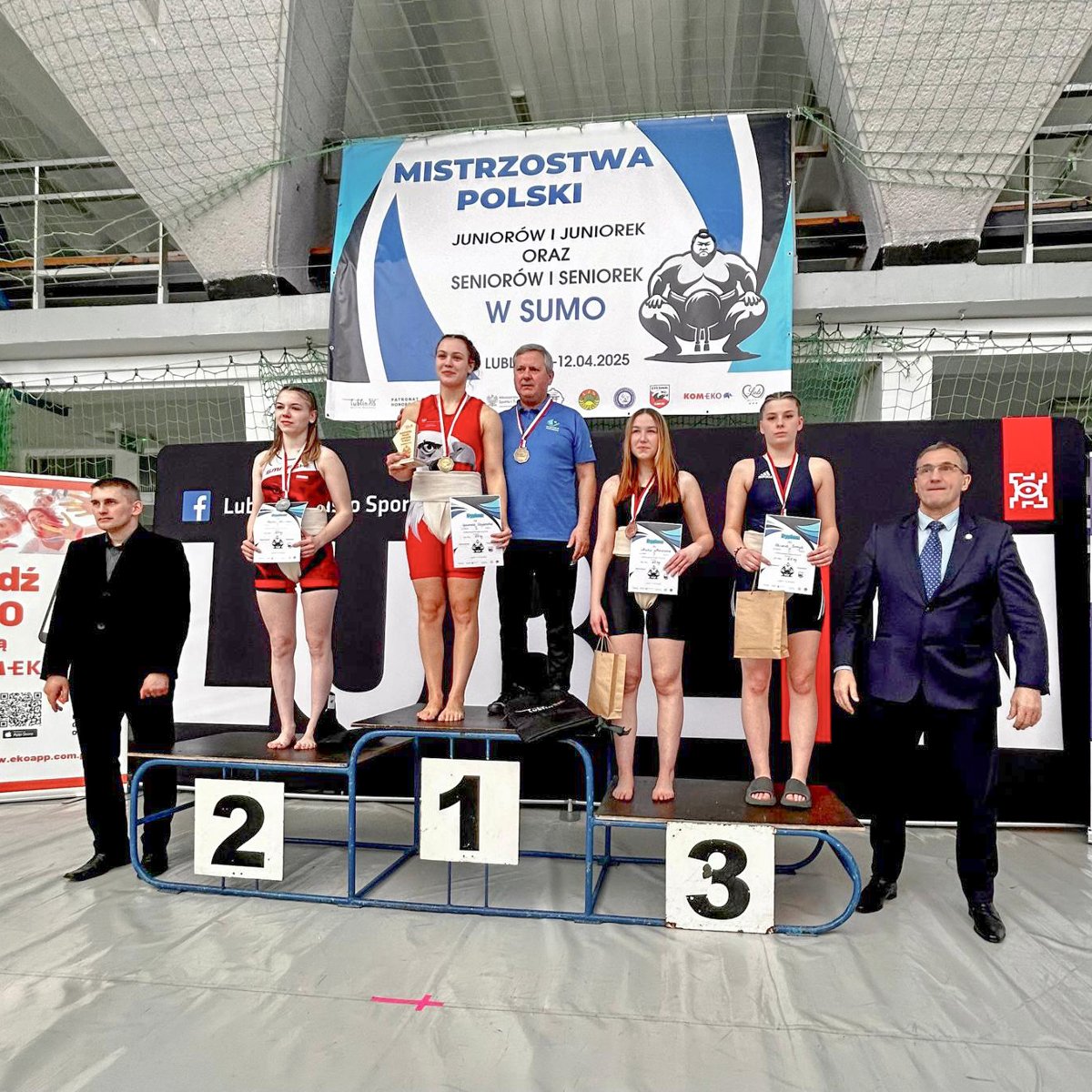 Zawodnicy Sobieskiego na MP seniorów i juniorów w sumo, fot. Jakub Tim Zawodnicy Sobieskiego na MP seniorów i juniorów w sumo, fot. Jakub Tim - na podium stoją 4 zawodniczki, a obok zwyciężyczni również jej trener. Po bokach 2 wręczających nagrody. W tle ścianka sponsorska oraz plakat zawodów