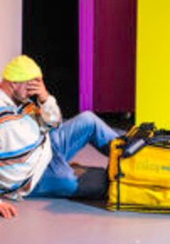 na zdjęciu mężczyzna siedzi pod ścianą, ręką zasłania twarz, przed nim na podłodze leży żółta torba termiczna do przewozu jedzenia