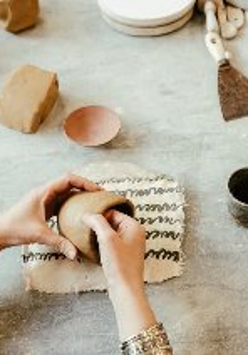 Osoba formuje naczynie z gliny na stole roboczym, na którym leżą różne narzędzia i kawałki gliny.