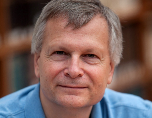 On a dark background, a smiling man with short gray hair. He is wearing a blue shirt.