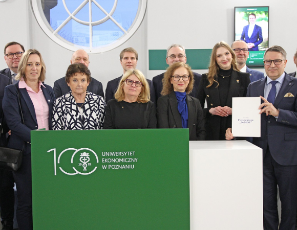 Grupa kilkunastu osób w strojach formalnych stoi za ladą z logo Uniwersytetu Ekonomicznego w Poznaniu; jedna z osób trzyma dokument porozumienia.