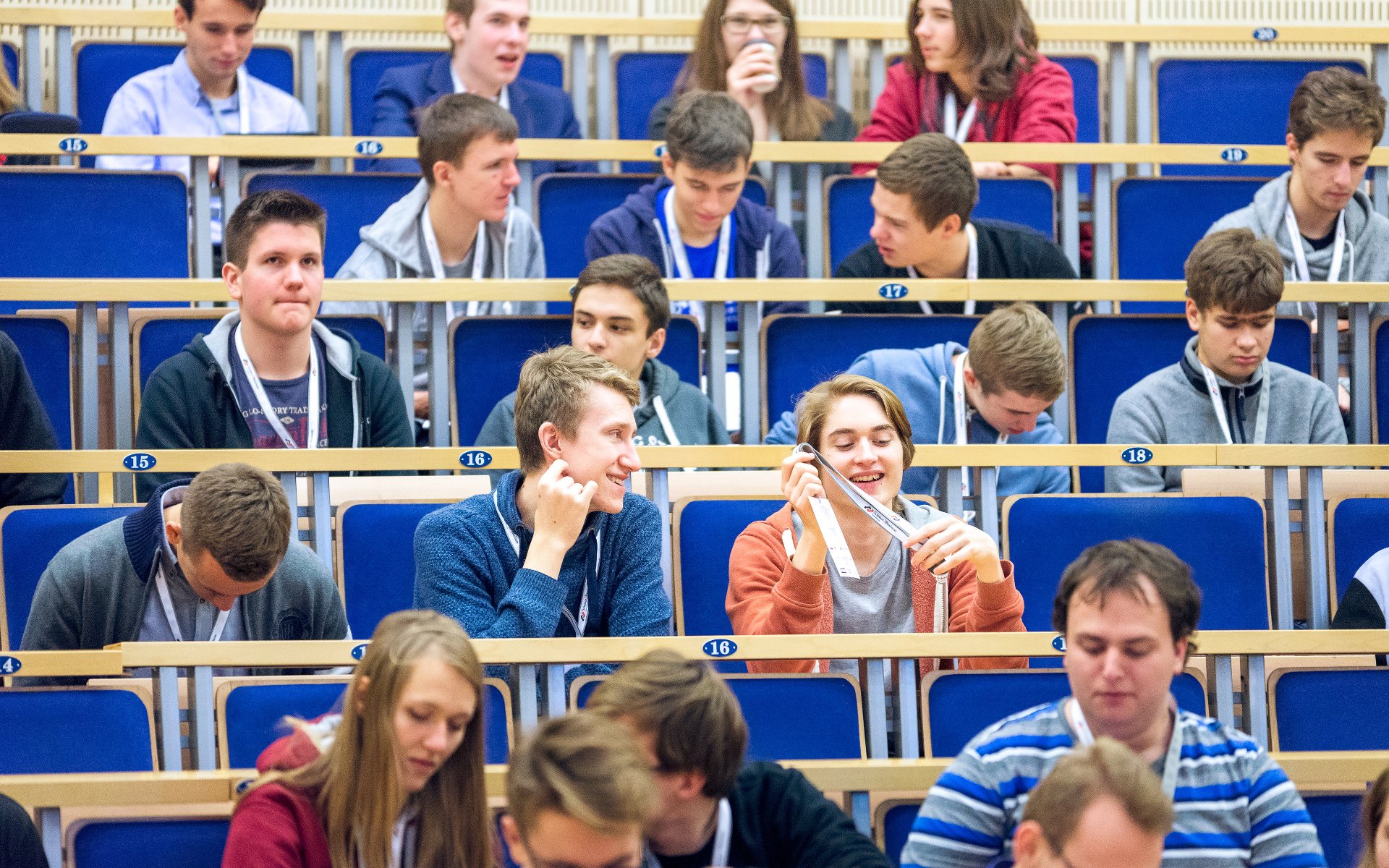 Grupa młodych ludzi siedzi na widowni sali wykładowej z niebieskimi krzesłami; mają identyfikatory i rozmawiają przed rozpoczęciem wydarzenia.