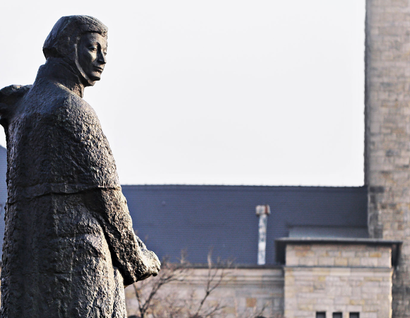 Kamienna lub metalowa rzeźba przedstawiająca stojącą postać w płaszczu, zwróconą bokiem i patrzącą w stronę fotografa. W tle widoczne są jasne, kamienne budynki i dachy, a całość ma spokojny, zimowy nastrój.