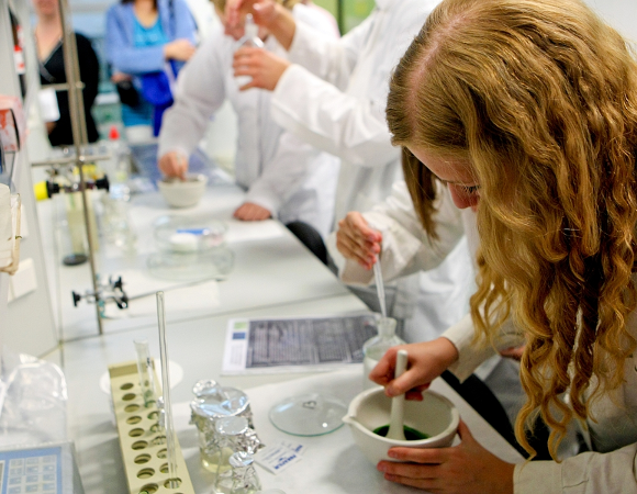 Kobieta w długach falowanych blond włosach w białym kitlu pochyla się nad stołem w laboratorium, miesza zielony płyn w małej białej misce. W tle niewyraźnie widać inne osoby