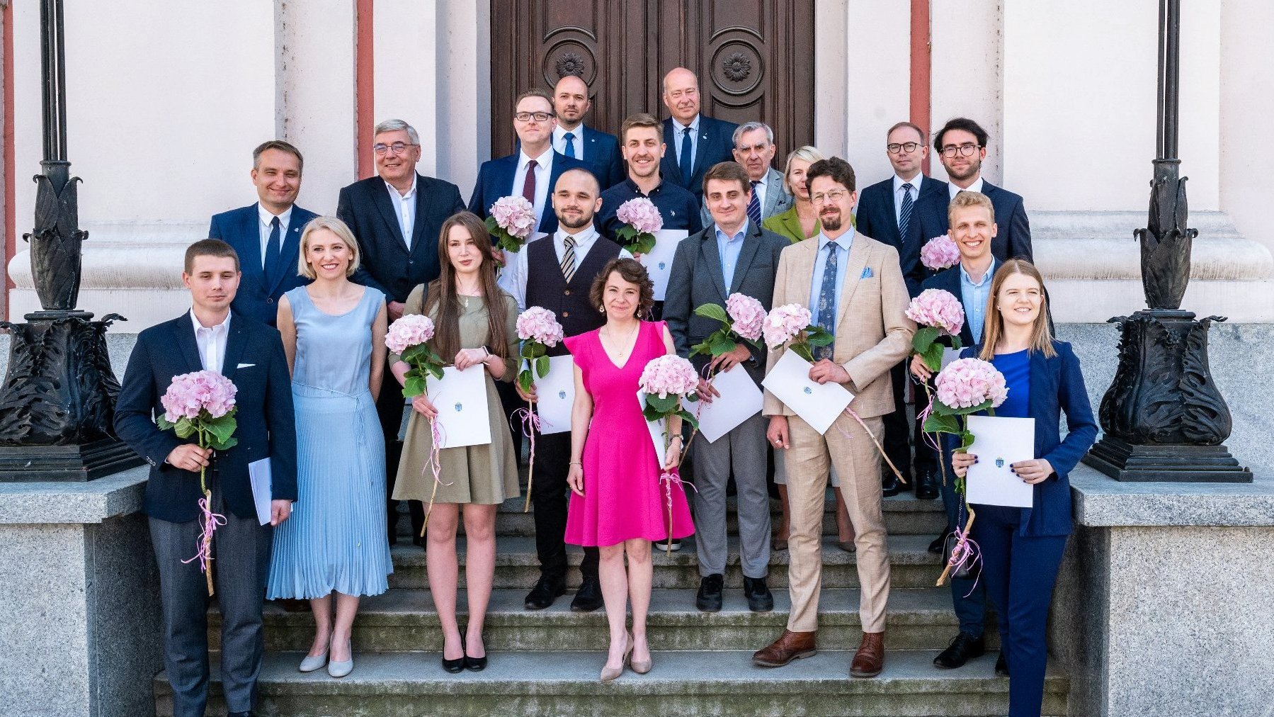 Zdjęcie przedstawia grupę elegancko ubranych osób stojących na schodach Urzędu Miasta Poznania. Część osób trzyma w rękach kwiaty i dyplomy.