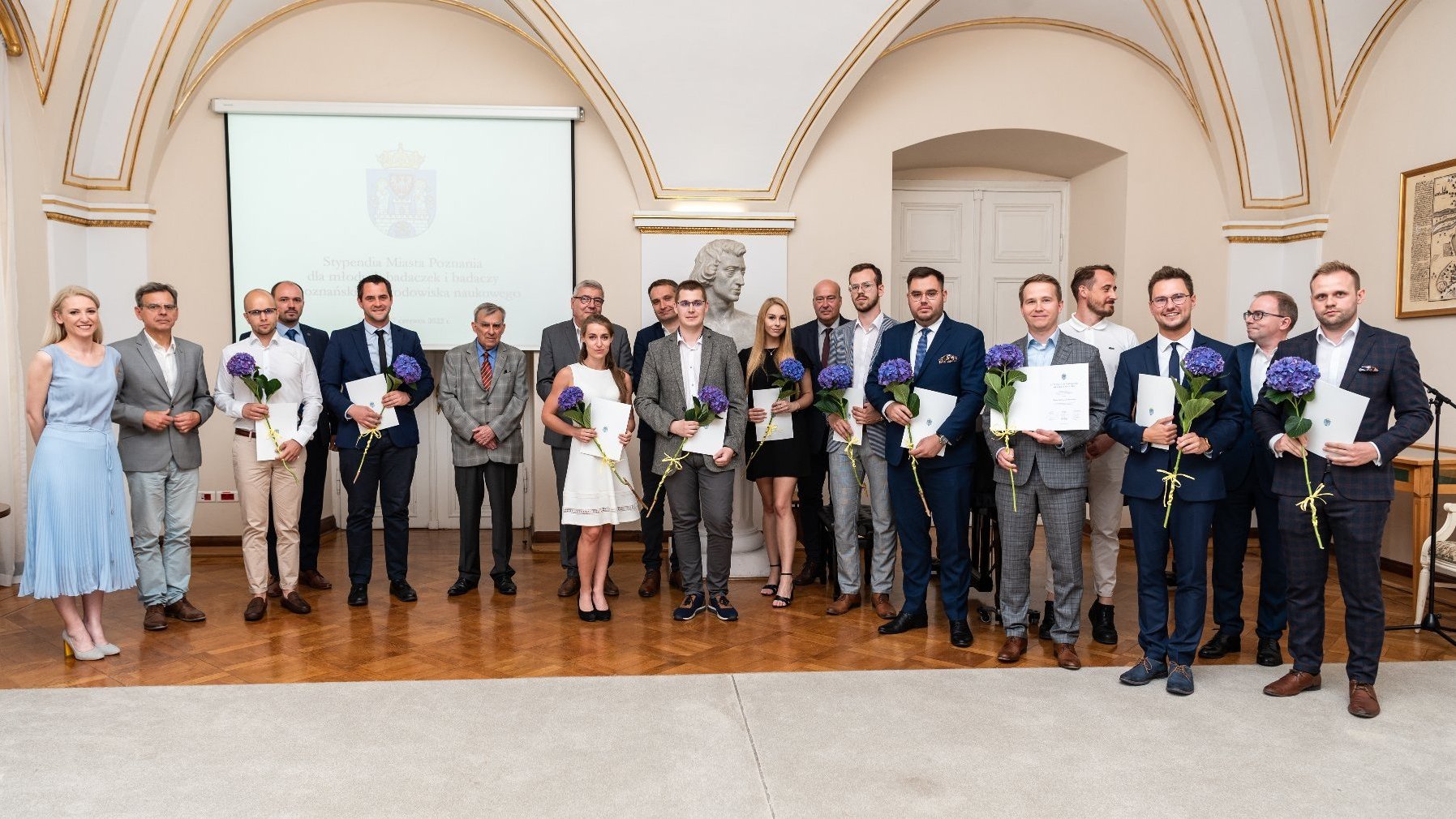 Zdjęcie grupowe laureatek i laureatów wraz z władzami Miasta i uczelni. Osoby stoją na tle slajdu tytułowego wydarzenia, w Sali Białej Urzędu Miasta Poznania.