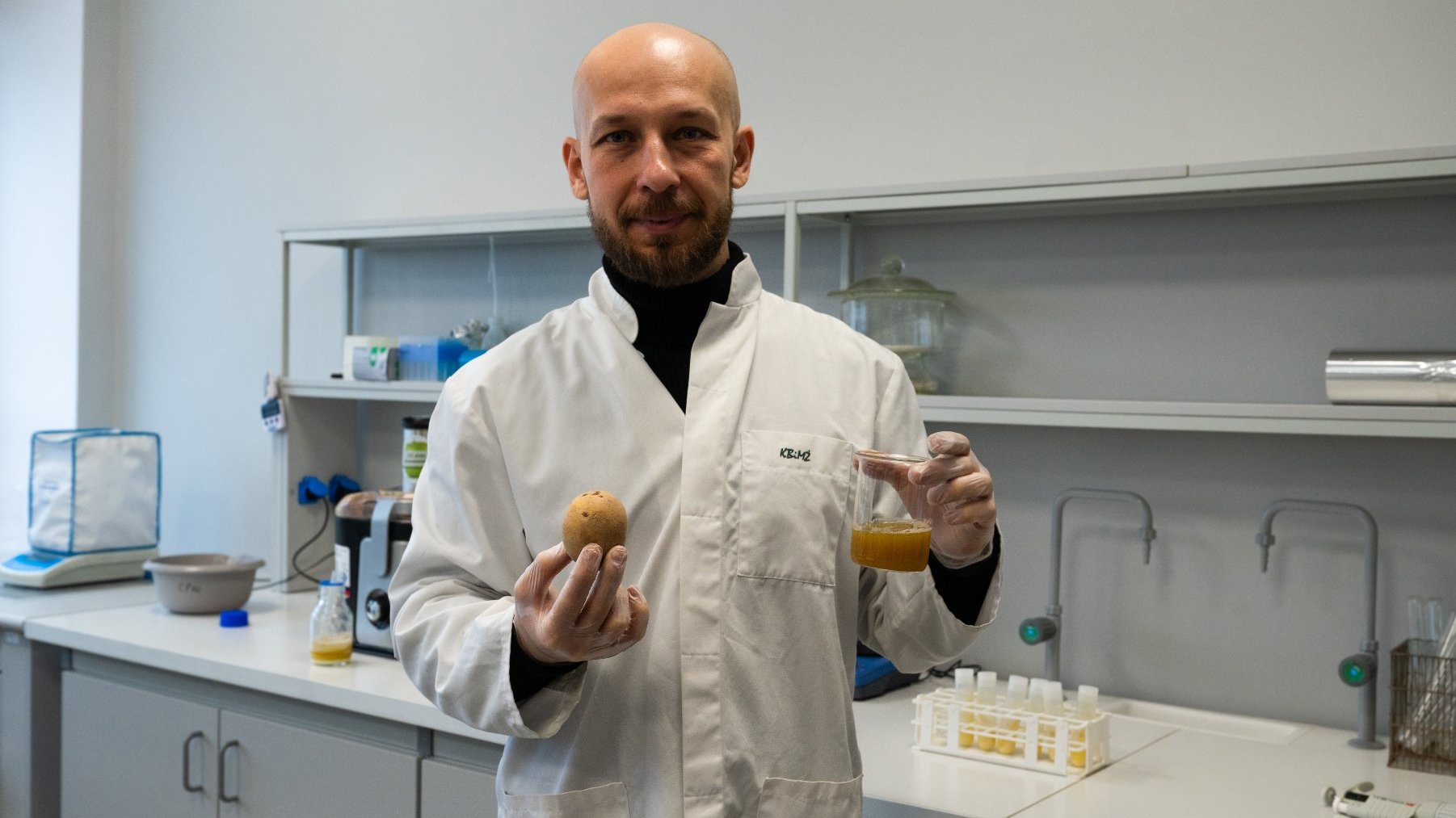 Zdjęcie przedstawia naukowca stojącego we wnętrzu laboratorium. Naukowiec jest w białym kitlu i trzyma w prawej ręce ziemniaka, a w lewej probówkę z sokiem z ziemniaka - grafika artykułu