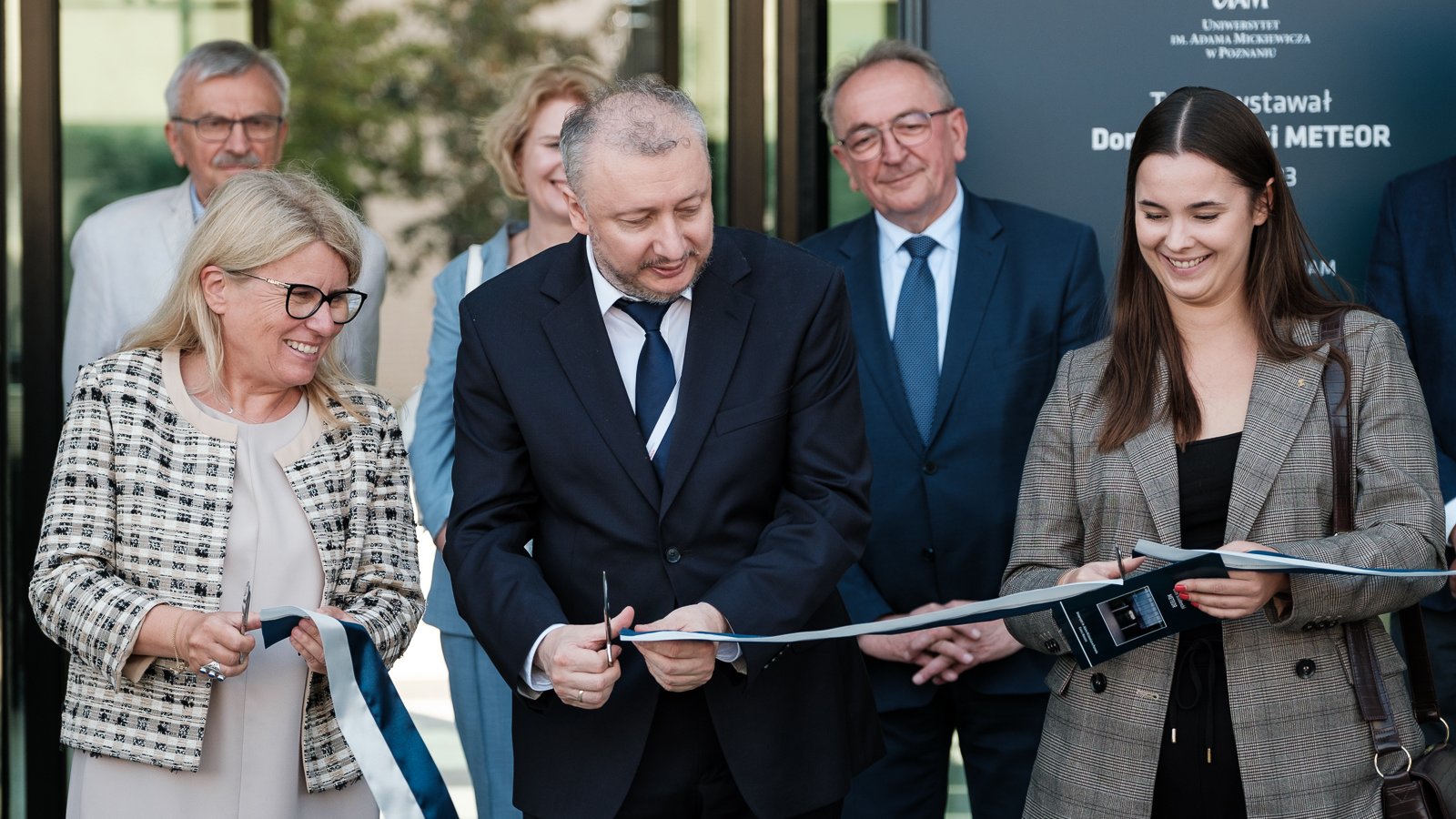 zdjęcie przedstawia trzy elegancko ubrane roześmiane osoby, w tym dwie kobiety i w środku mężczyznę, przecinających wstęgę