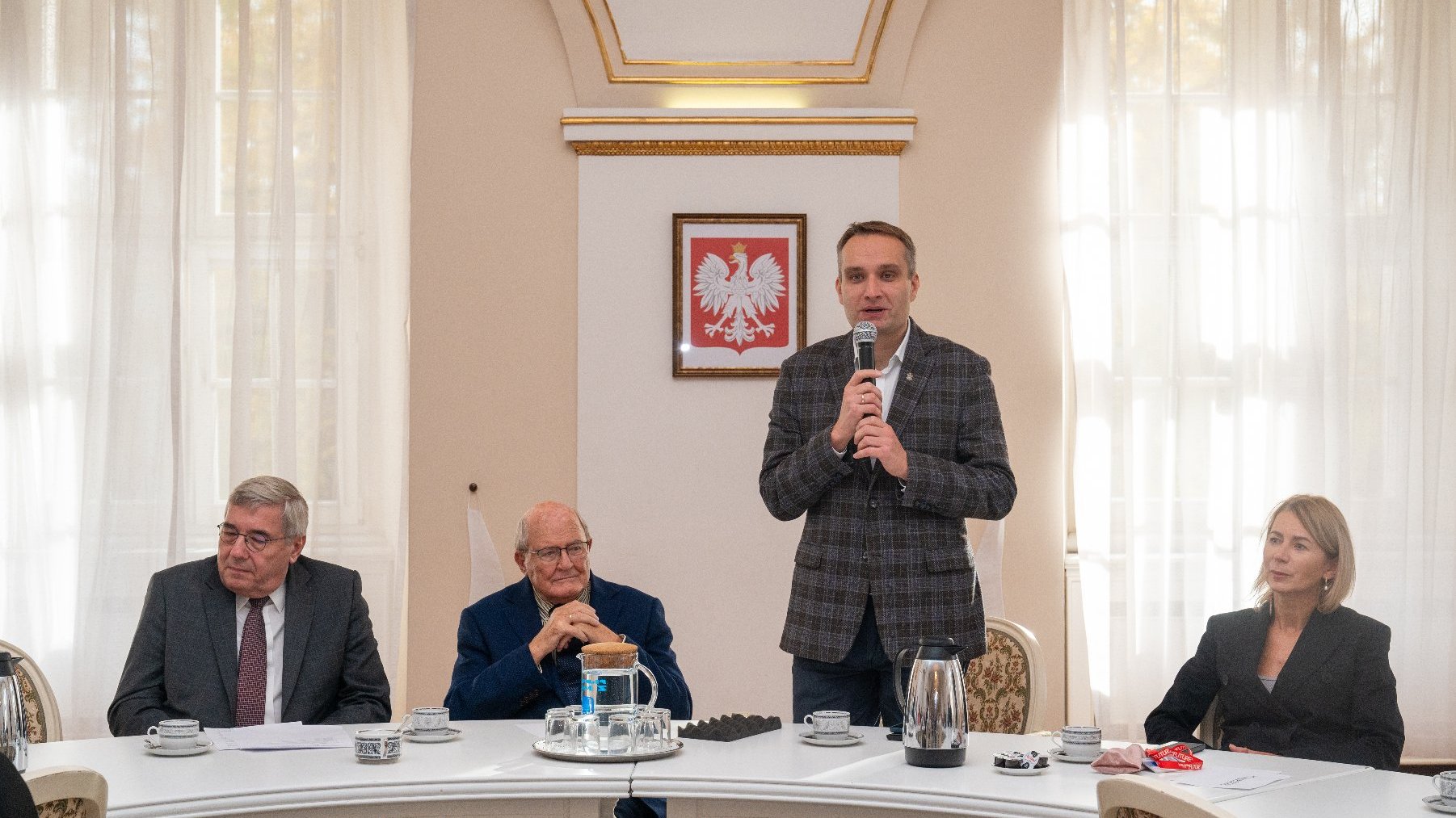 w dużej sali przy stole siedzi jedna kobieta i dwóch mężczyzn, jeden mężczyzna stoi i mówi przez mikrofon