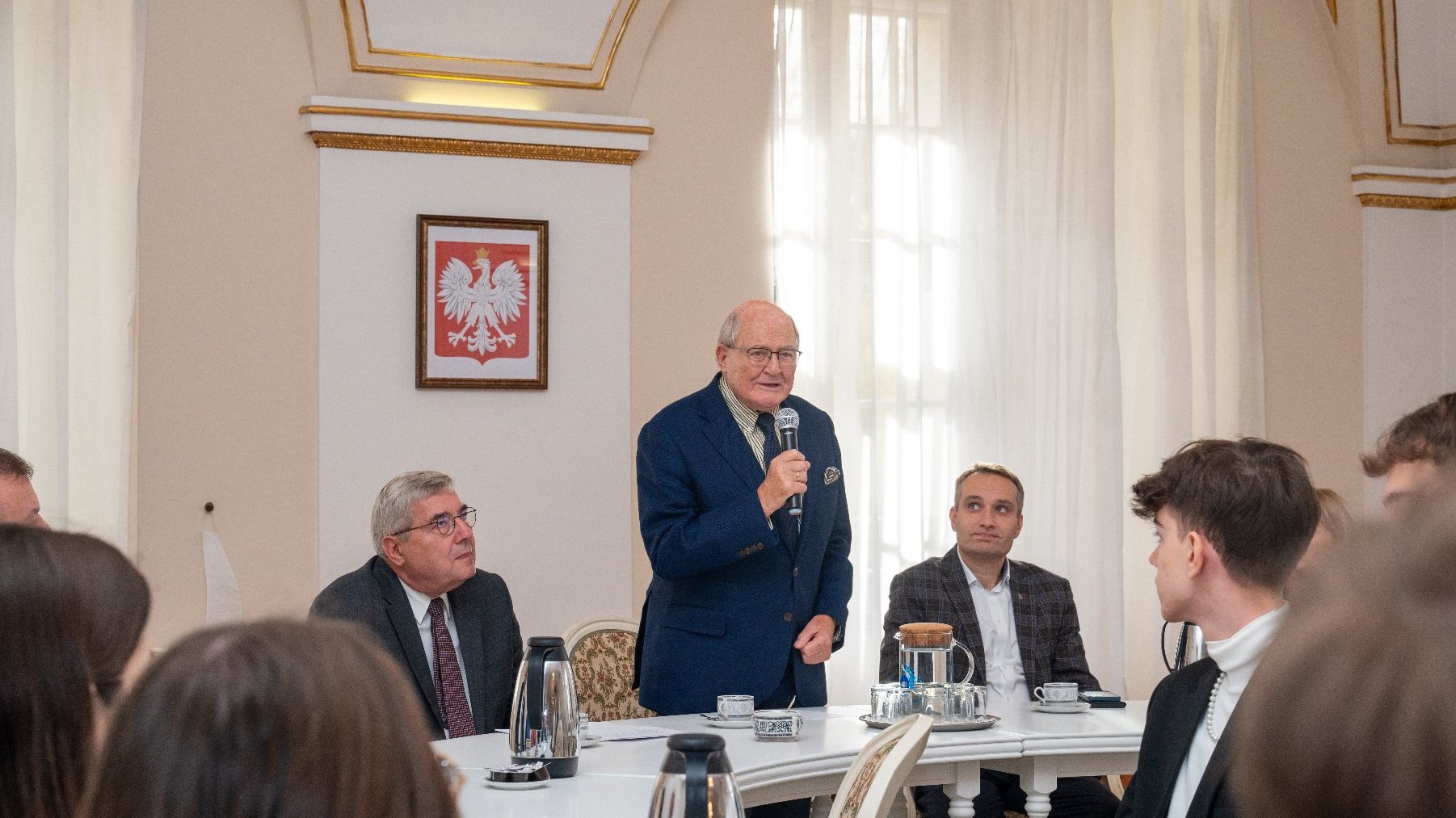 w dużej sali przy stole siedzi parę osób i patrzą na mężczyznę który stoi i mówi przez mikrofon