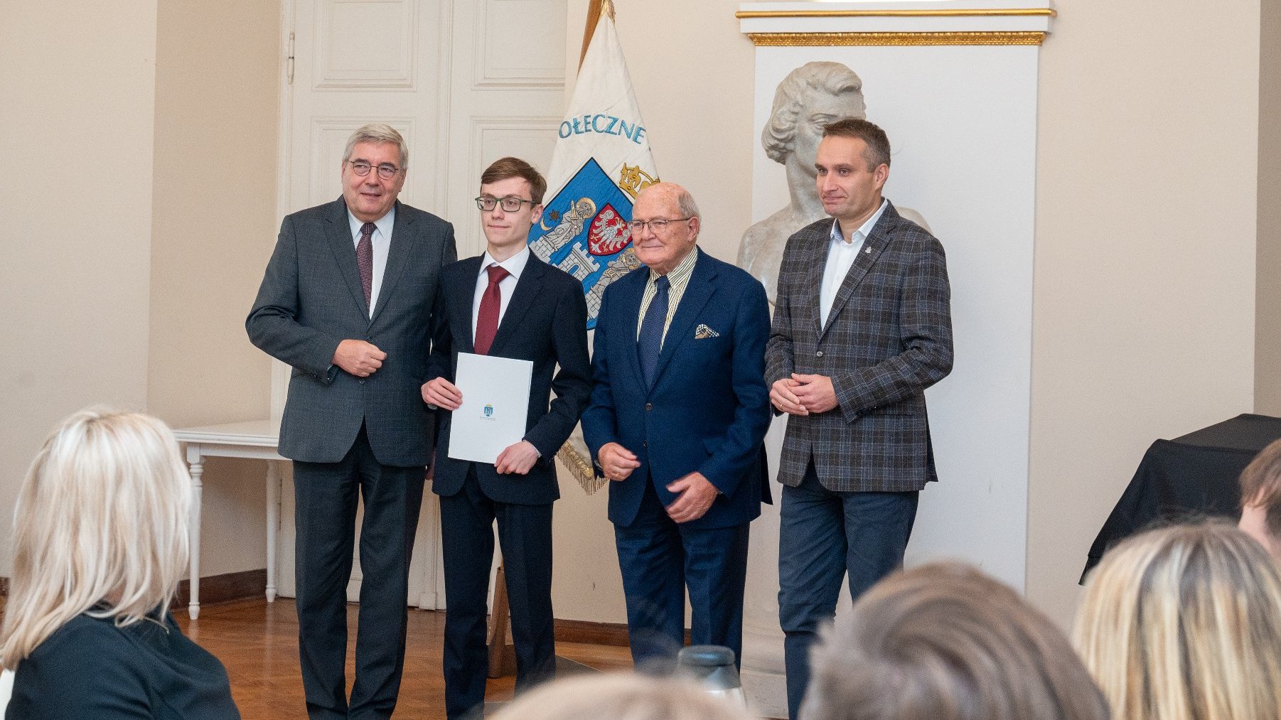 w sali stoi 4 mężczyzn, jeden z nich trzyma dyplom