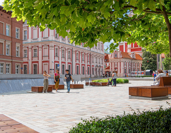 Słoneczny plac miejski z ławkami i fontanną, otoczony zabytkowymi budynkami; spacerują ludzie, a nad placem rozpościerają się zielone korony drzew