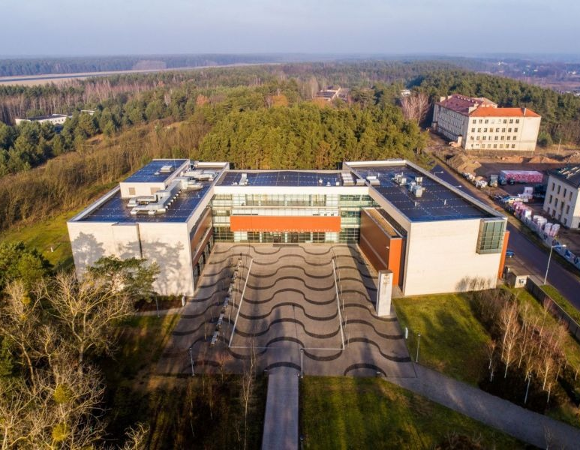 Widok z lotu ptaka na nowoczesny, duży budynek w kształcie litery U, otoczony lasem. Przed wejściem znajduje się plac z falistym wzorem nawierzchni, a wokół widać drzewa i kilka innych budynków.