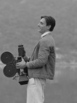 Czarno-białe zdjęcie mężczyzny stojacego bokiem do obiektywu i trzymającego kamerę na tle zamglonego krajobrazu.