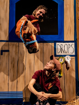 Sena z przedstawienia: przy drewnianej ścianie siedzi kobieta, która patrzy na konia (pacynkę) wystawiajacego łeb przez otwór w ścianie.