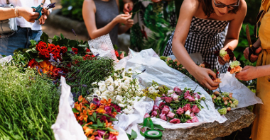 Na zdjęciu stół z roślinami i kobiety plecące wianki