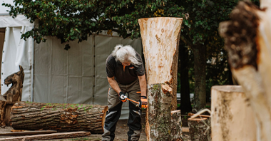 Na zdjęciu mężczyzna z dłutem pochyla się przy rzeźbionym drewnie.