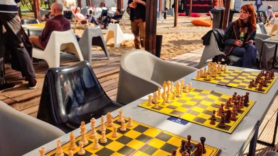 Żółte szachy na szarych stołach. Stoliki z krzesałami stoją na drewnianych platformach na plaży. Sporadycznie na krzesłach siedzą ludzie. W tle leżaki i plaża.