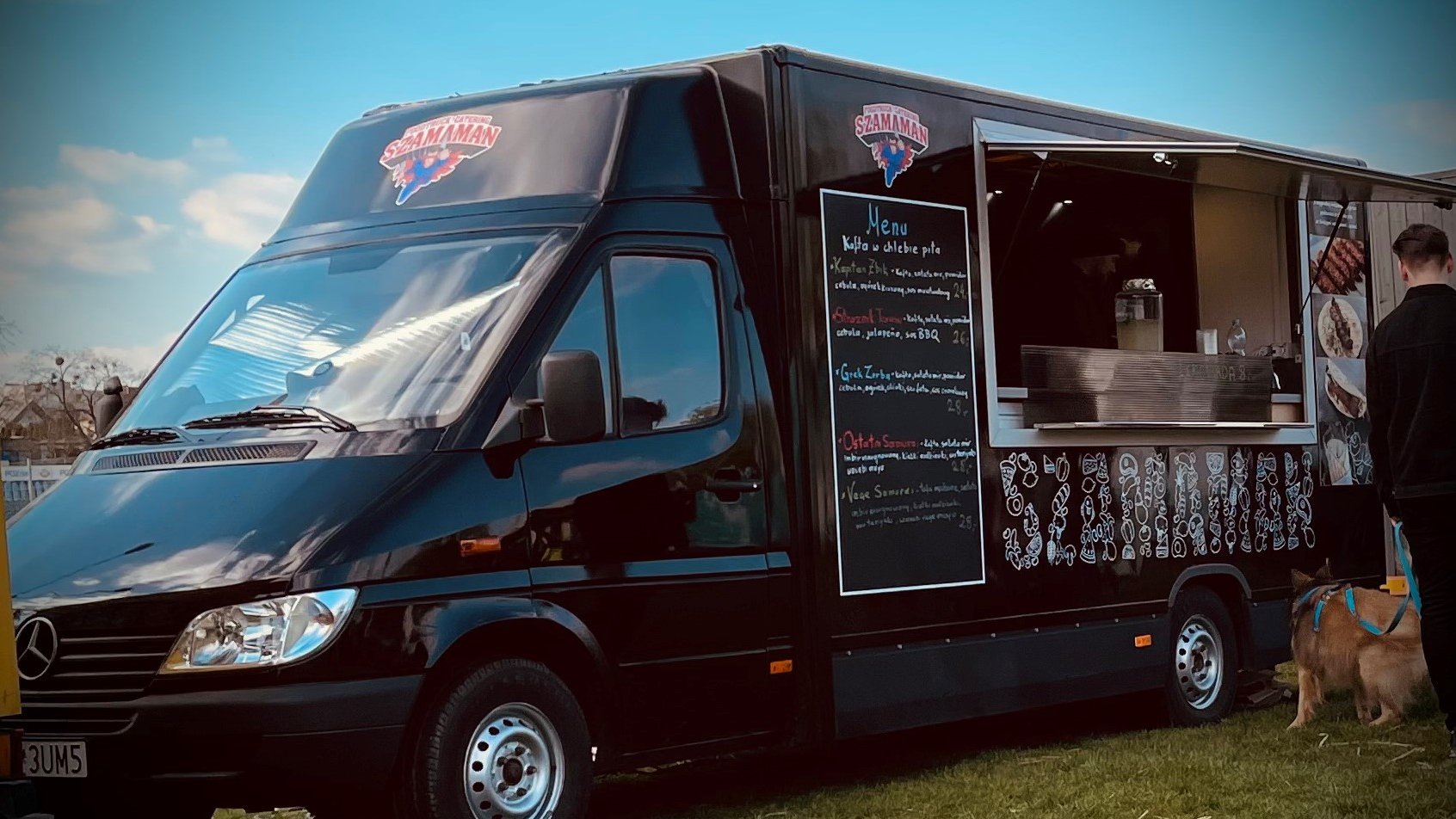 Czarny food truck Szama-Man stoi na dworze. Pod okienkiem stoi mężczyzna z psem. Na tablicy wypisane Menu.