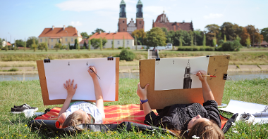 Mama z dzieckiem leżą na kocu na trawie i rysują Katedrę Poznańską widzianą z drugiego brzegu rzeki Warty