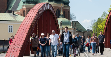 Przewodnik prowadzi dużą grupę zwiedzających przez most, w tle Katedra Poznańska