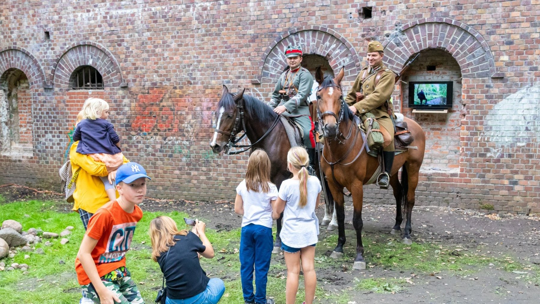 Dwaj mężczyźni siedzą na koniach ubrani w wojskowe mundury. Przed nimi grupa dzieci (jedno z nich robi zdjęcie), za nimi mury fortu.