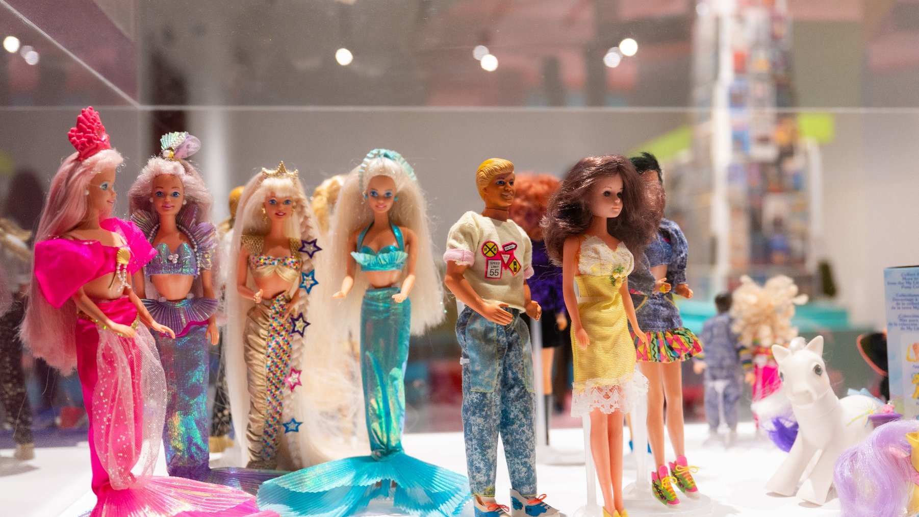 Zdjęcie przedstawia gablotę z lalkami Barbie ubranymi w kolorowe sukienki różnej długości. Wśród nich stoi Ken ubrany w dżinsy i sportową bluzę.