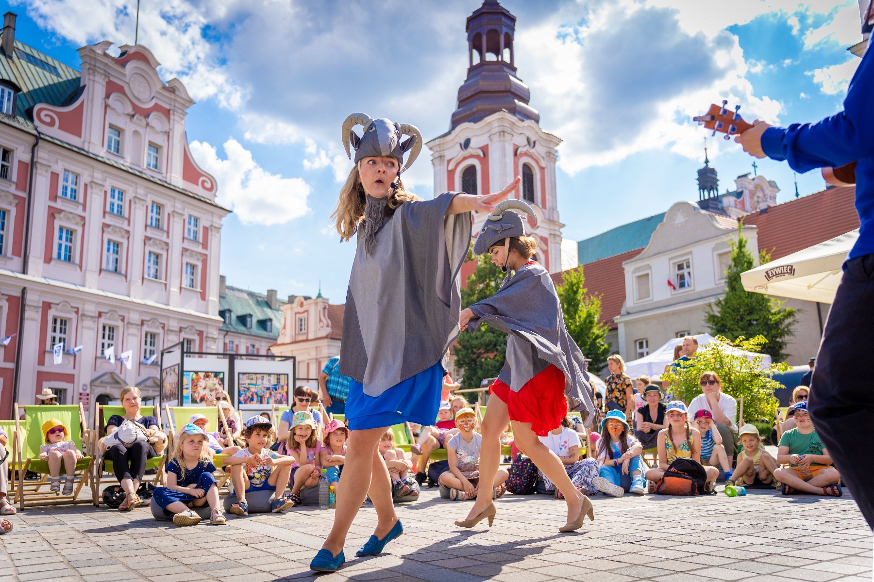 Wielkimi krokami zbliżają się imieniny Piotra i Pawła, patronów Poznania. Zdjęcie przedstawia dwie kobiety przebrane za koziołki oraz tłum patrzących na nie dzieci. W tle widac budynek urzędu miasta.
