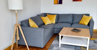 Na zdjęciu wnętrze mieszkania, widać szarą kanapę z żółtymi poduszkami, na ścianie dwa obrazy, obok stół i lampa