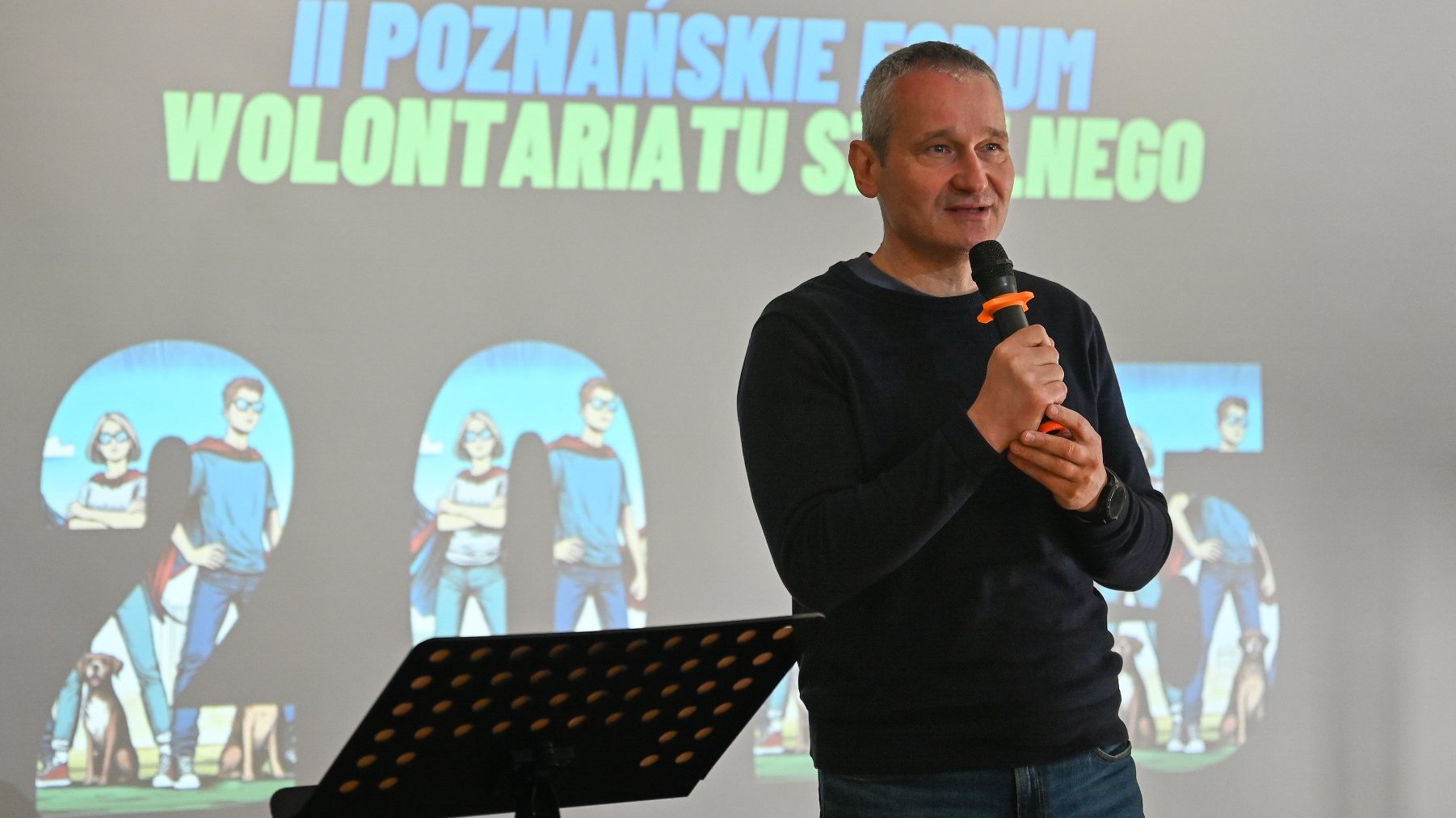 Na zdjęciu mężczyzna w ciemnym swetrze, mówiący coś do mikrofonu