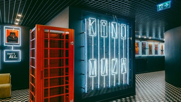 It's the interior of a cinema with a modern retro style. A large neon sign reading "KINO MUZA" glows on the wall. Next to it stands a red British-style phone booth. Illuminated movie posters hang on the walls, and the floor features a black-and-white checkerboard pattern. Dark tones dominate the space, accented by bright neon lights, creating a moody, artistic atmosphere.